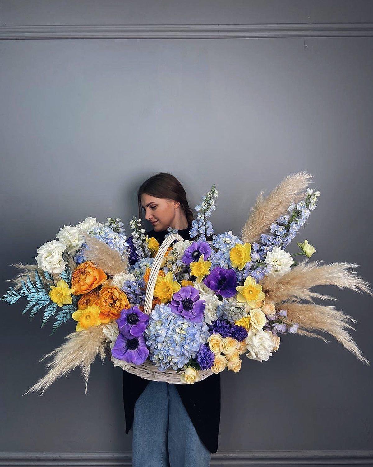 Florist holding massive spring basket with purple anemones, orange roses, blue hydrangea, and pampas grass