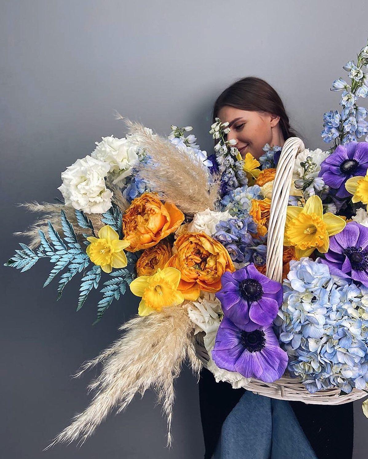 Florist holding vibrant spring basket with daffodils, anemones, orange roses, and blue delphinium