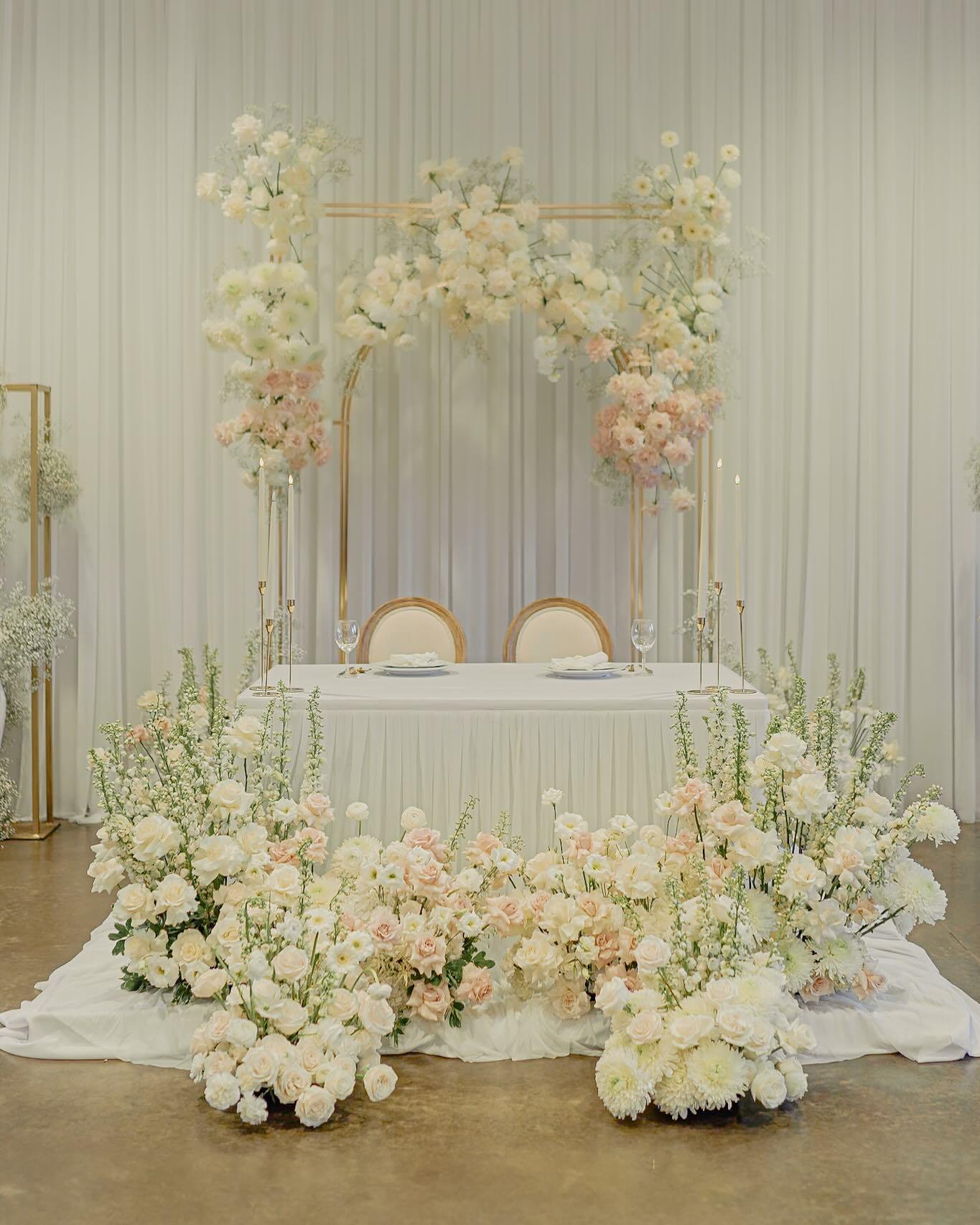 Elegant sweetheart table with white and blush floral arch, gold accents, and abundant roses at wedding reception
